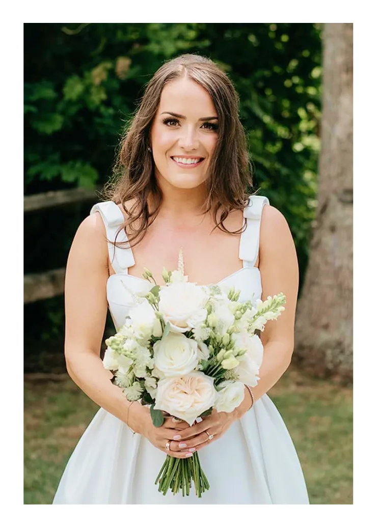 Smiling bride in white dress holding a bouquet of white and pale pink flowers outdoors.