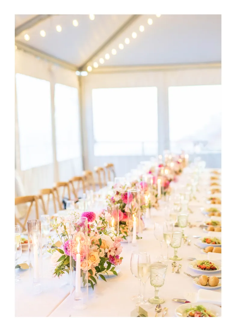 Long table with floral centerpieces, candles, and place settings in a bright, tented event space.