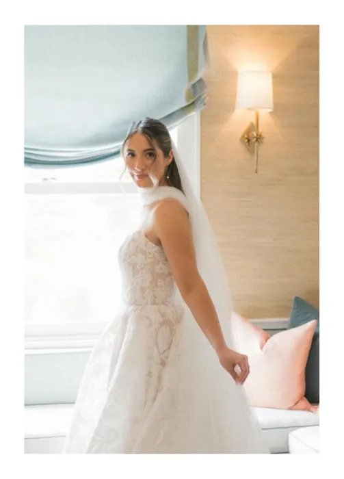 Bride in white lace wedding gown and veil standing indoors near a window and sofa.