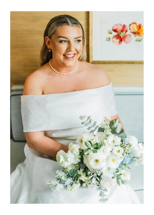 Smiling bride in off-shoulder white dress holding a large bouquet of white flowers seated on a couch.