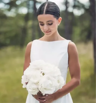 Bride in white dress holding a bouquet of white flowers outdoors with a blurred forest background.