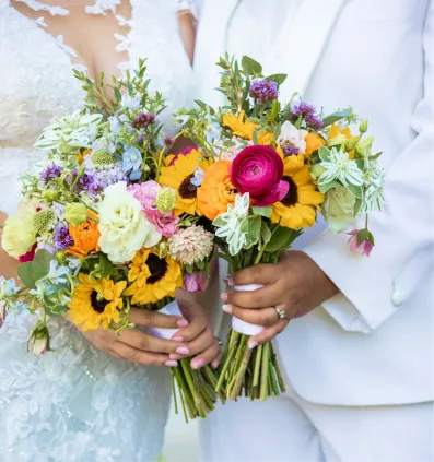 Two people in wedding attire each holding colorful bridal bouquets with sunflowers and ranunculus.