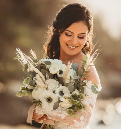 Smiling bride with dark hair holding a bouquet of white flowers and greenery outdoors.