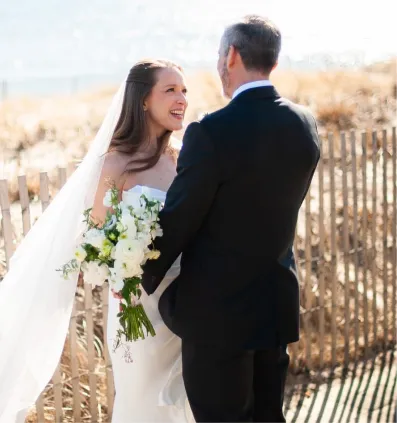 Bride smiling while holding white flower bouquet facing groom in black suit near wooden fence outdoors.