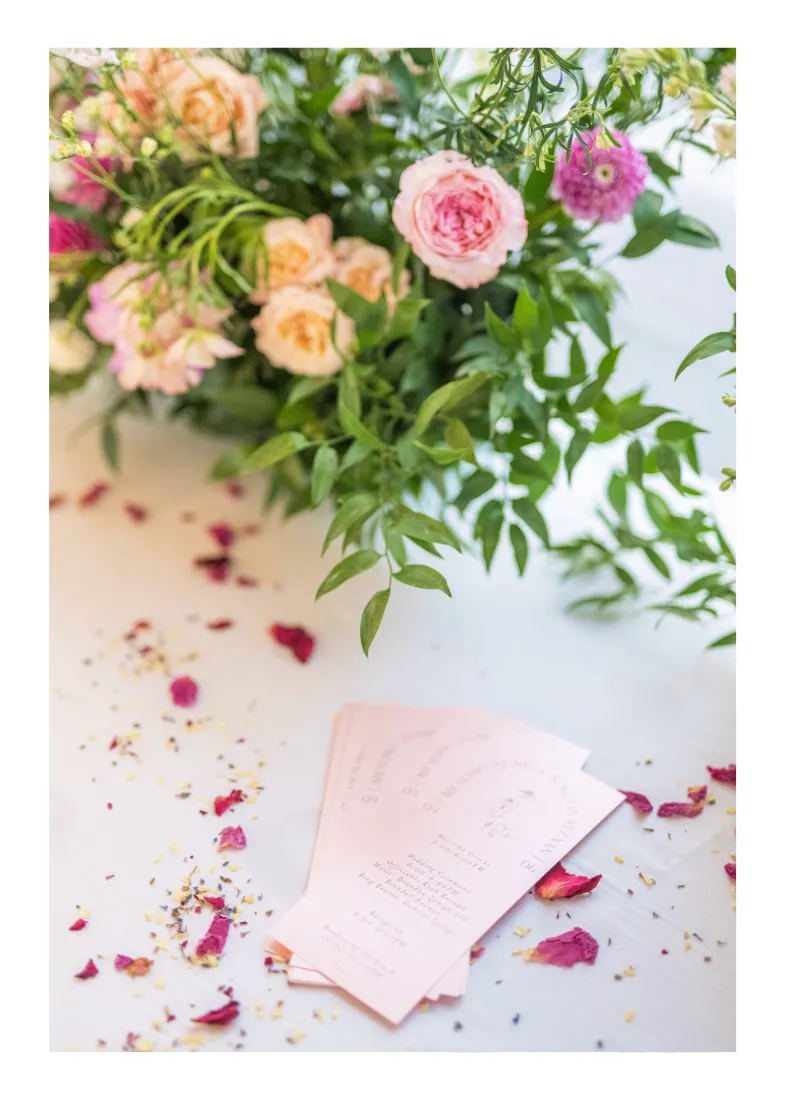 Pink and peach roses with greenery next to pink wedding ceremony programs and scattered petals.