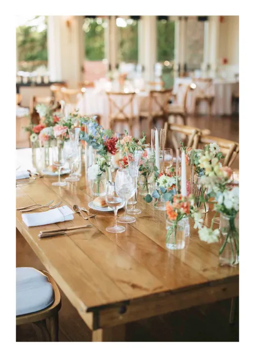 Wooden table set with floral arrangements, glassware, plates, and cutlery in a bright event space.
