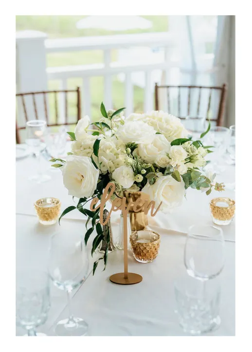 White floral bouquet centerpiece with greenery and table number 'eleven' on a white tablecloth.