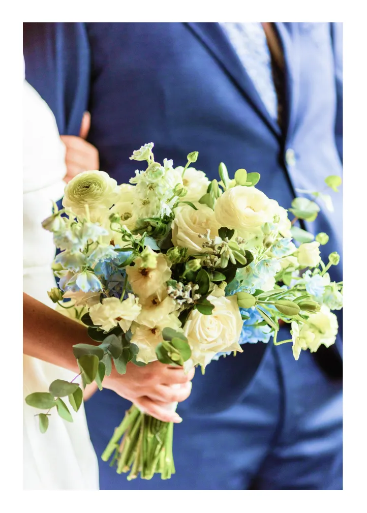 Bride holding a bouquet of white and pastel flowers with green leaves, groom in a navy suit nearby.
