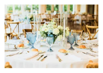 Elegant wedding table setup with white floral centerpiece, blue glasses, bread rolls, and silver cutlery.