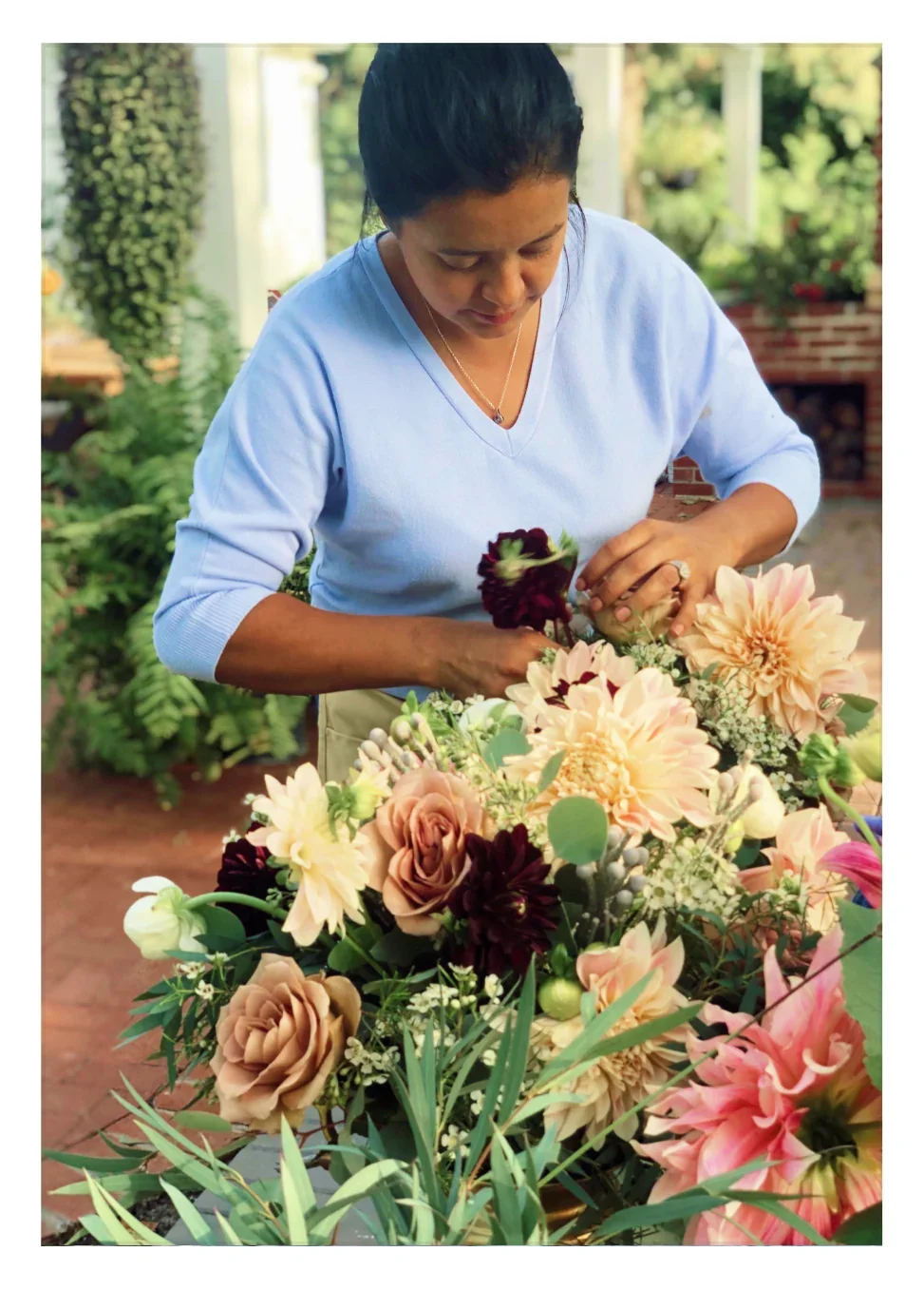 Smiling woman in white blouse holding a small gift box, standing in front of blue and purple flowers.