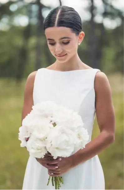 Bride in white dress holding a bouquet of white flowers, looking down with a soft smile.