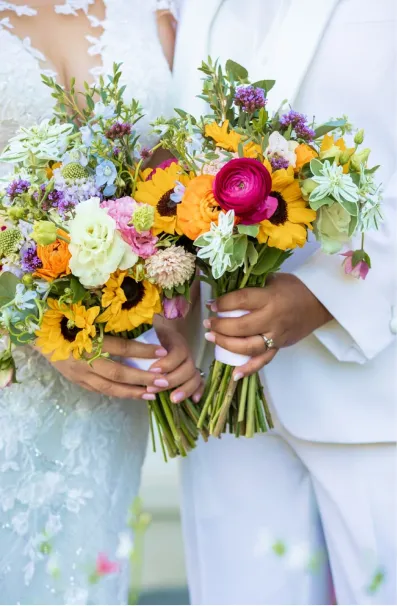 Two people in white attire holding colorful flower bouquets with sunflowers and various blooms.