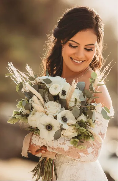 Smiling bride in lace dress holding a bouquet of white flowers and greenery outdoors.