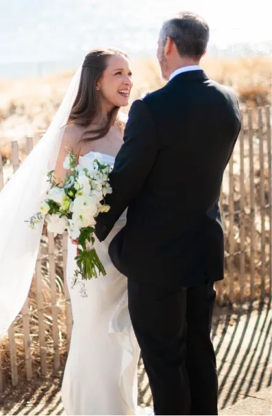 Bride in white dress and veil holding bouquet, smiling at groom in black suit outdoors.