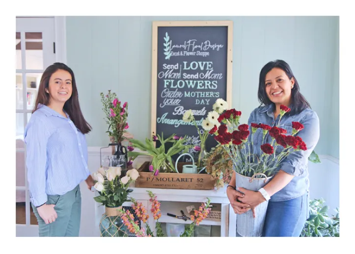 Two women smiling in a flower shop with assorted flowers and a chalkboard sign about Mother's Day.