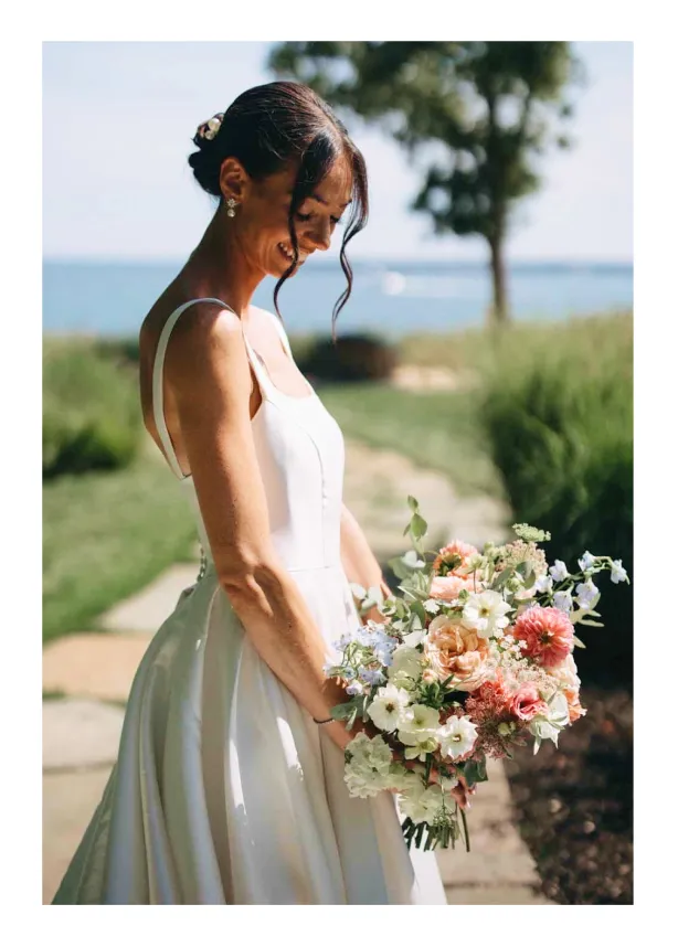 Smiling bride in white dress holding a colorful bouquet outdoors with greenery and water in background.